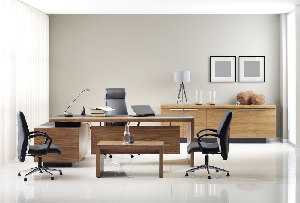 This image depicts a workspace with a modern, minimalist design. It features a wide desk accompanied by a sleek office chair, with a laptop, a notebook, and a cup placed on the desk. Behind the desk is a window letting in natural light, providing a bright and airy ambiance. The room's decor includes a stylish shelf with books and plants, adding a touch of green to the otherwise neutral palette. This image is perfect for illustrating a professional and clean office environment suitable for productivity and focus.